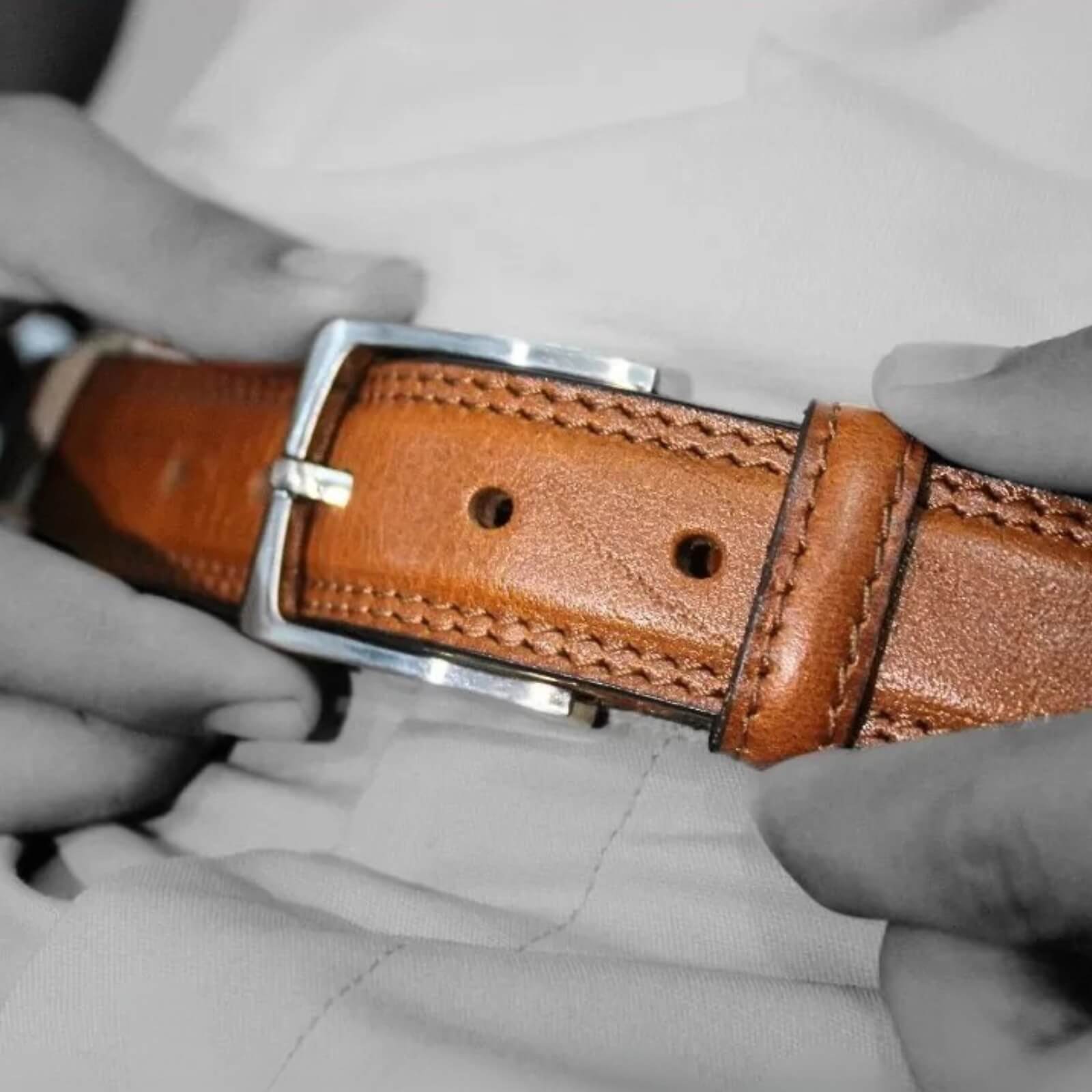 Brown leather belt with silver buckle held by gloved hands against a gray background